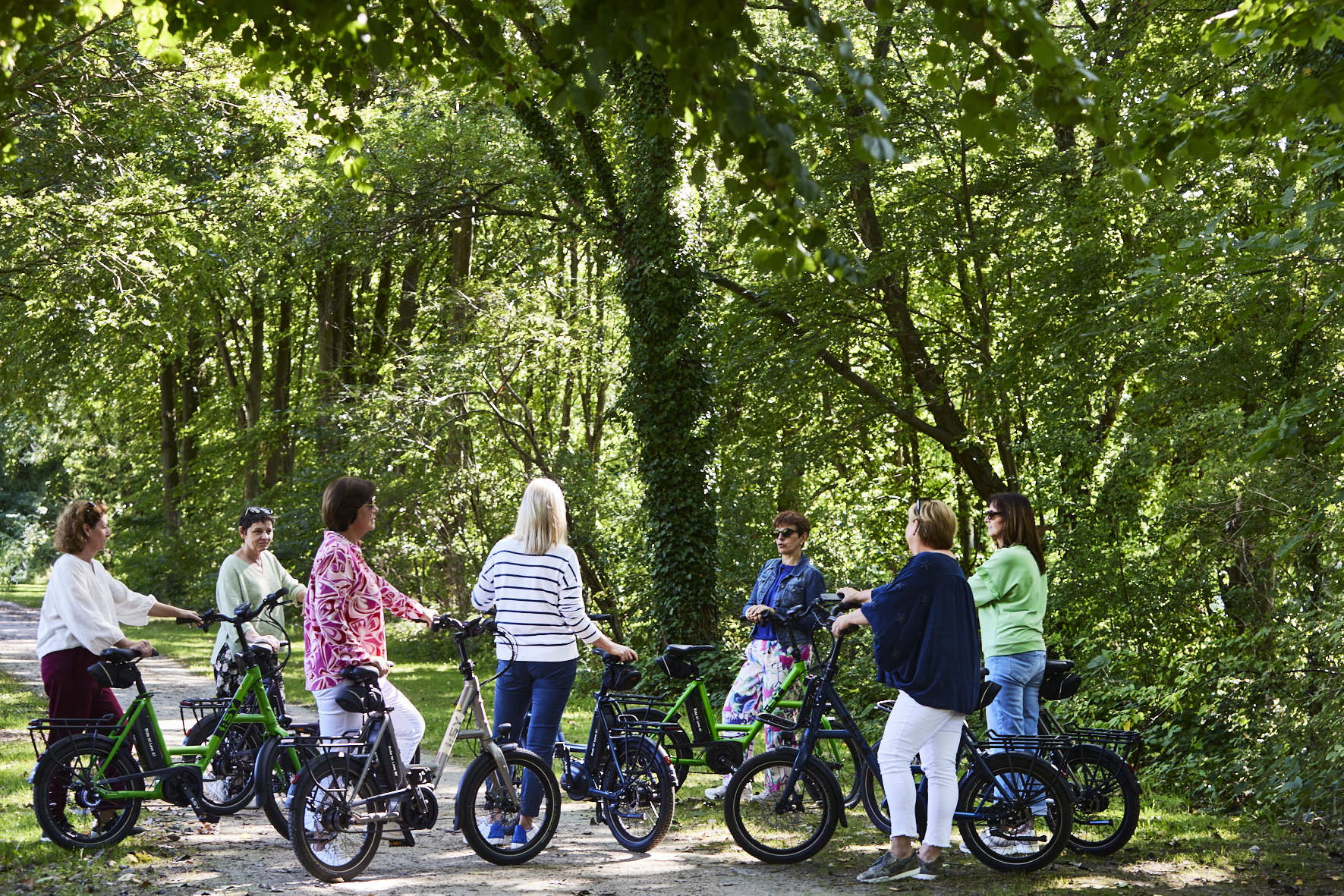 Groep mensen fietst samen in een groen park, met groene fietsen langs een pad onder bomen.