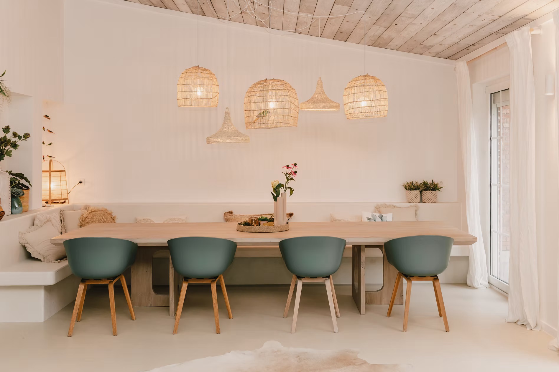 Witte eetkamer met houten tafel, vier groene stoelen, hanglampen van riet en bloemen op tafel.